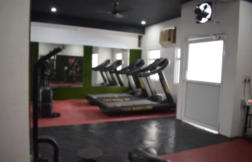 A row of treadmills beside a cable weight machine, facing a mirrored wall, with a wall-mounted air conditioning unit and a ceiling fan above.